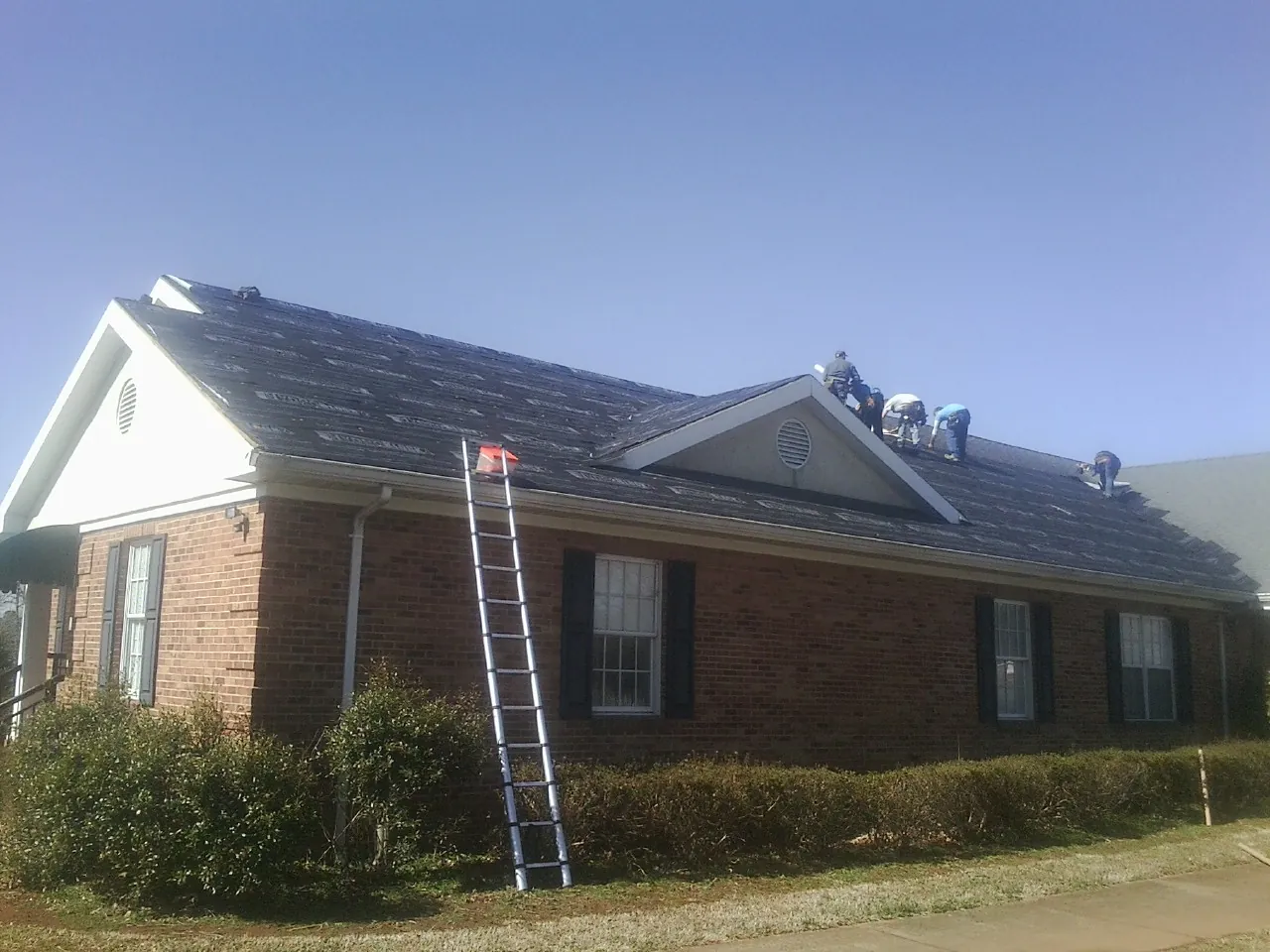 Crew tearing shingles off a brick ranch roof with a ladder set — Metal Roof Repair work by our crew in Saint Ann