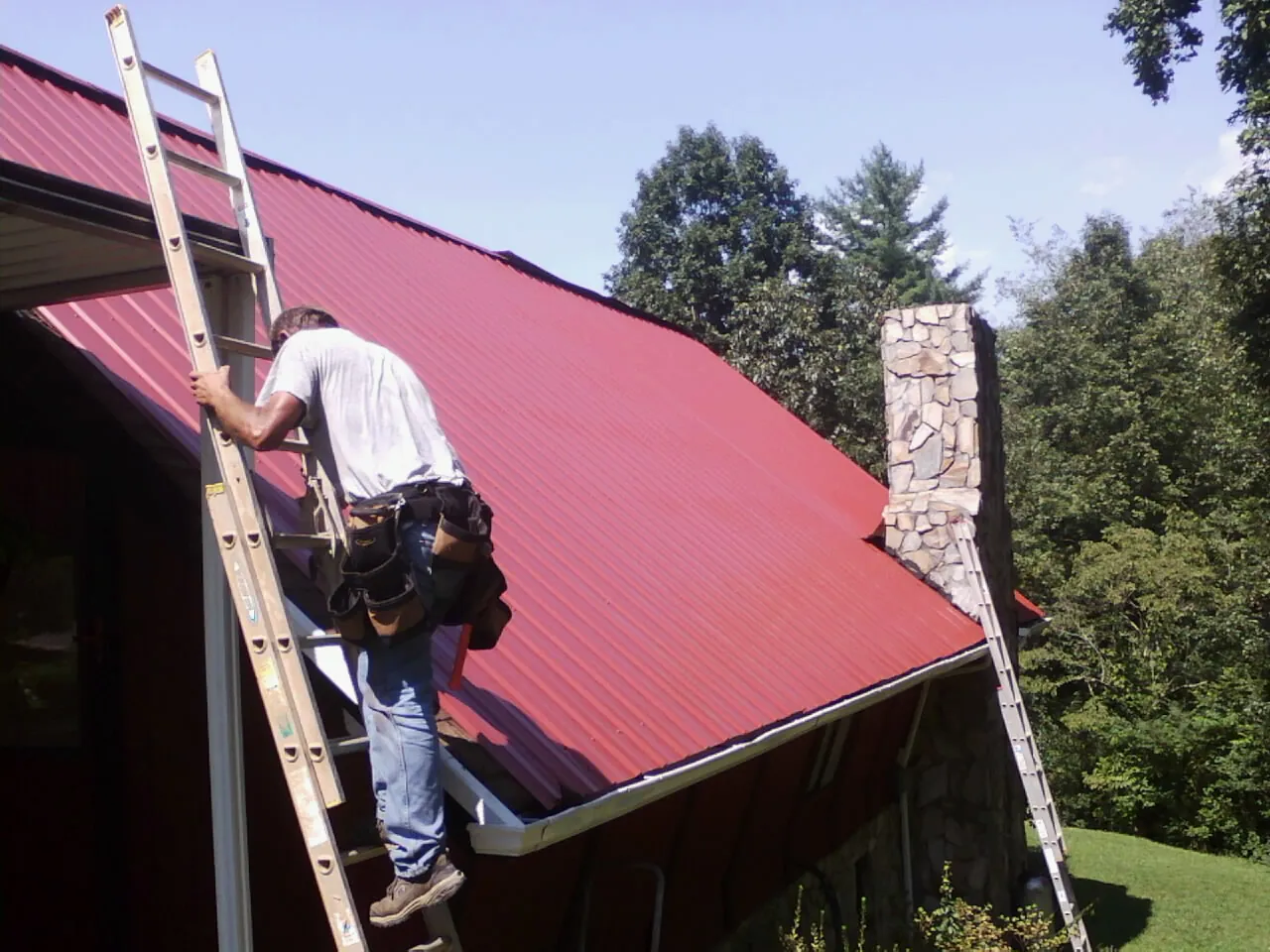 Roofer on a ladder beside a red metal roof and stone chimney — Soffit & Fascia Repair by Roofing Brothers in Saint Ann